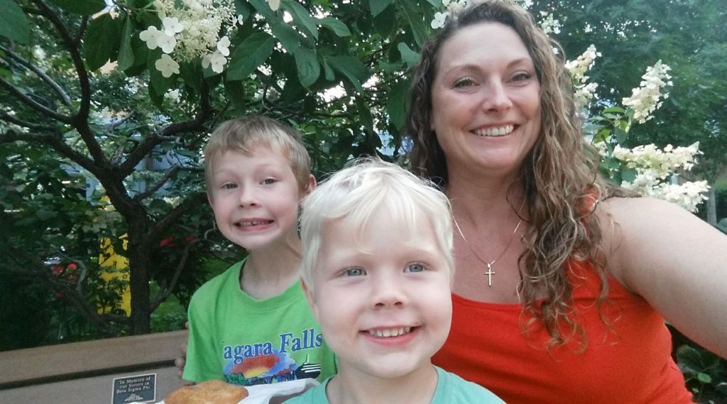 a smiling white woman with long curly brown hair takes a selfie with her two young boys on a park bench in kamloops bc